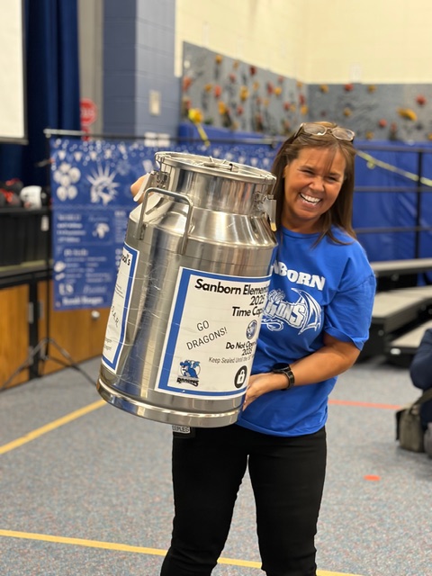 A smiling woman in a blue shirt holds a large silver milk can, standing in a school gym with banners in the background.