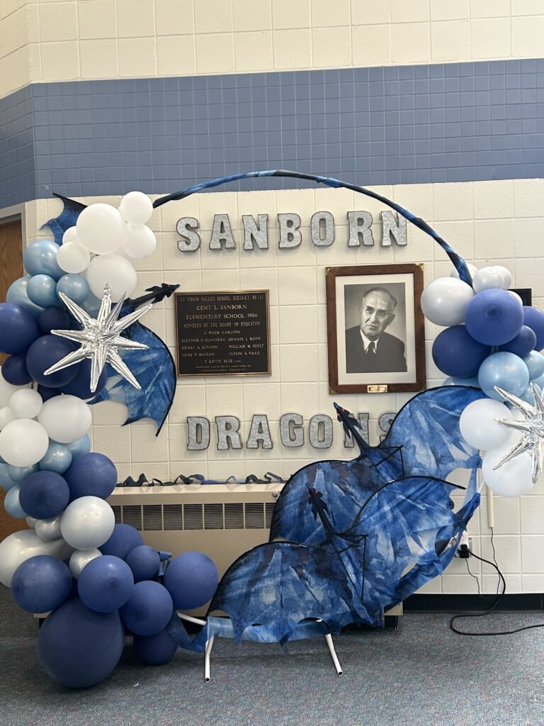A balloon arch in blue and white colors with a star and dragon decorations, featuring a portrait and "Sanborn Dragons" sign.