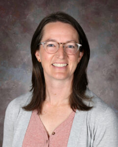 A woman with glasses and brown hair smiles, wearing a light gray cardigan over a pink top, against a textured background.