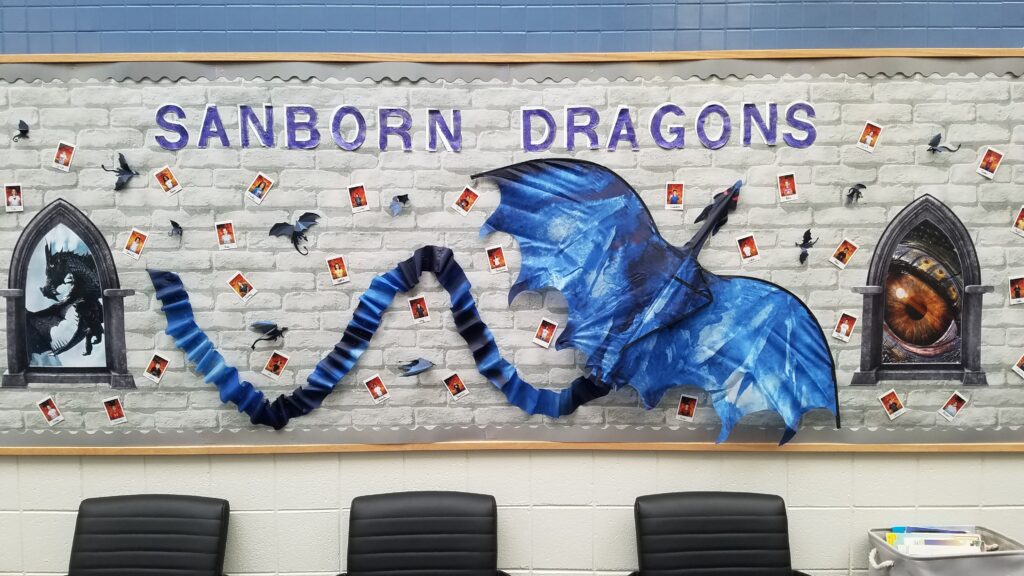 Bulletin board with a stone background.  There are 2 medieval looking window with a dragons eye peering out.
There is a blue dragon kite in the center with student pictures around it.  The wording is Sanborn Dragons.