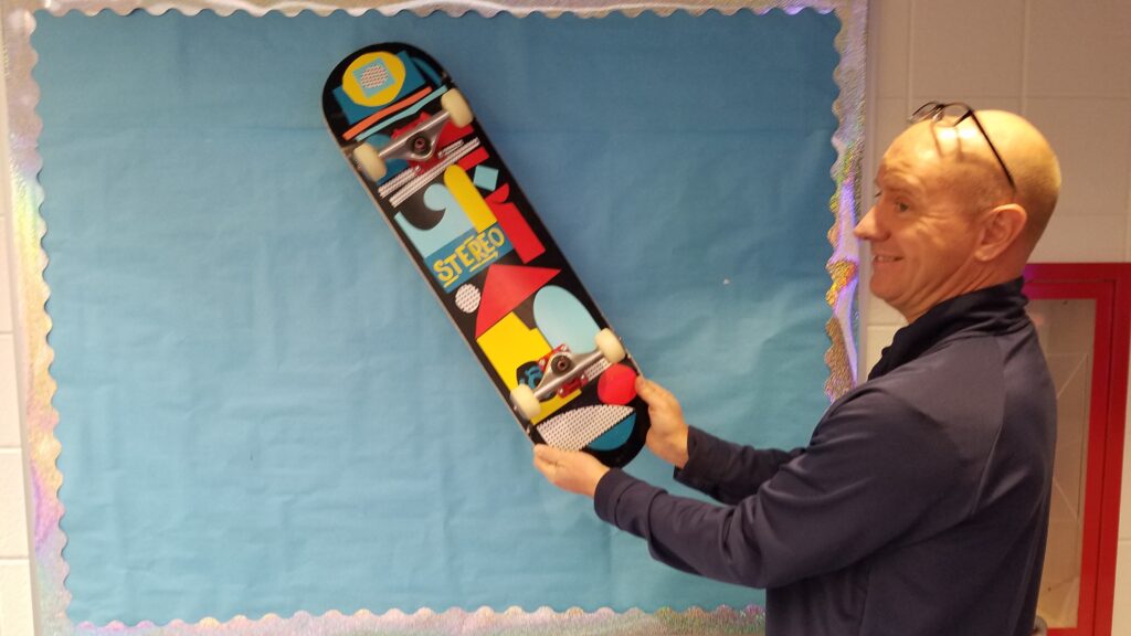 Photograph of a skateboard--black base with multicolored geometric shapes with the word STEREO on it being held by our PE teacher.