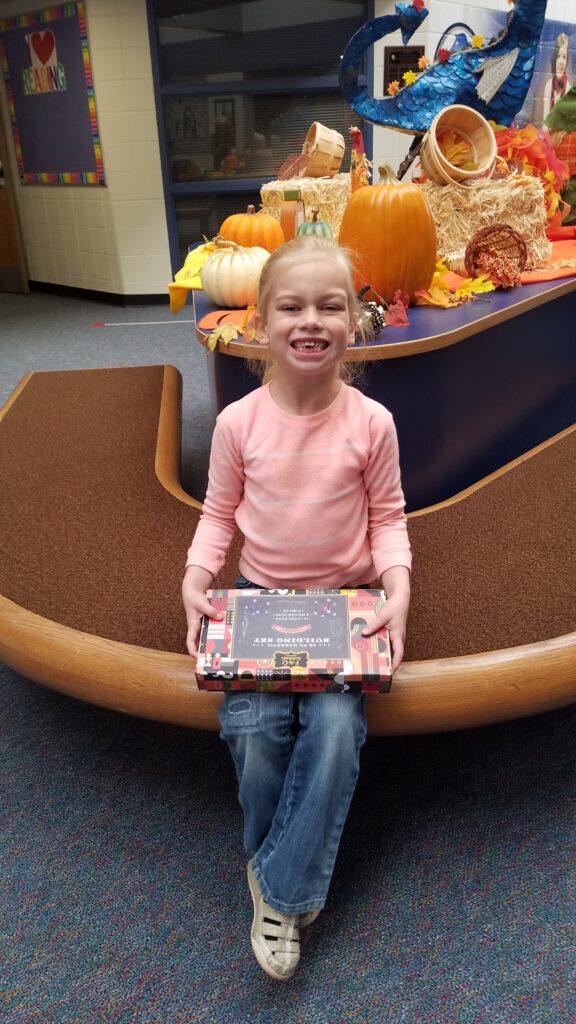 Photograph of a female student with blond hair, pink shirt and blue  holding a magnet building set.  She is sitting on a bench with fall decor behind him.