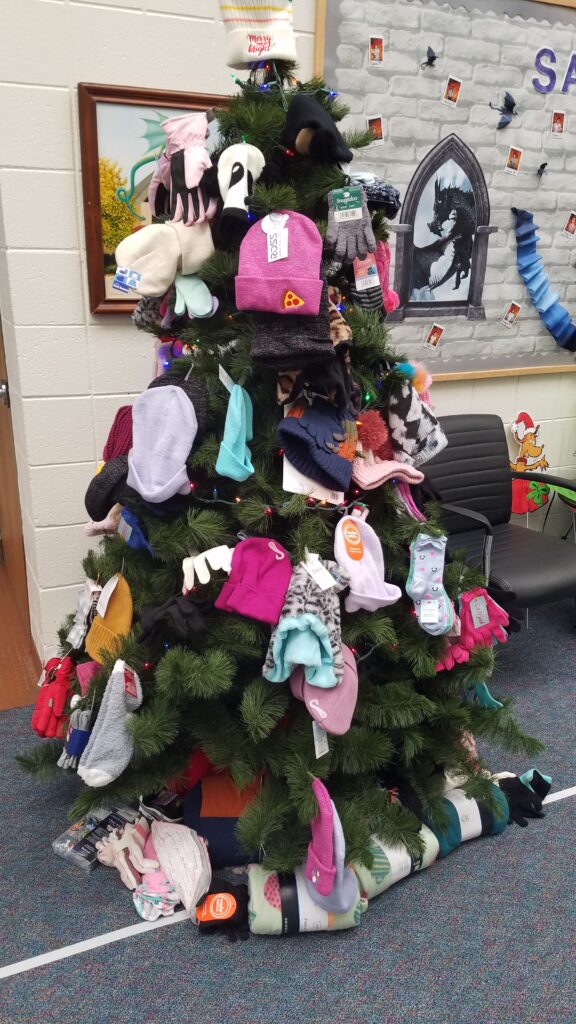 Lighted green artificial tree with mittens, socks, hats hanging on it.