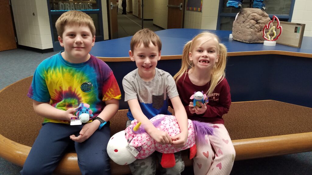 Photo of 3 students sitting on a bench.  Student #1 is a blond boy with a tie died shirt holding a small blue, stuffed dragon, student #2 is a boy with a gray t-shirt holding a pink & white stuffed dragon with white hearts, student #3 is a blond girl wearing a maroon shirt holding a blue stuffed dragon.