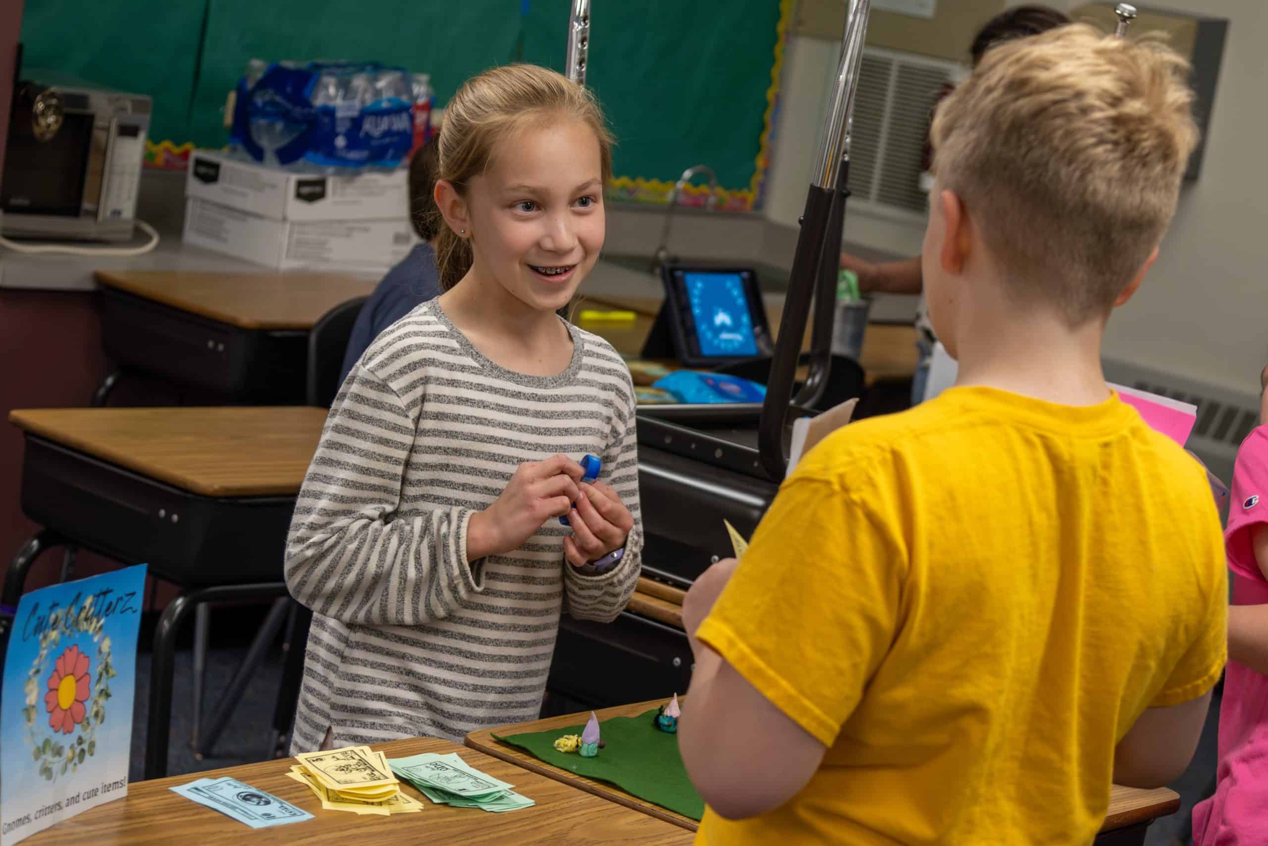 Female 5th grader standing behind desk holding a figure she made while trying to sell it to a male student in front of her at the class market.