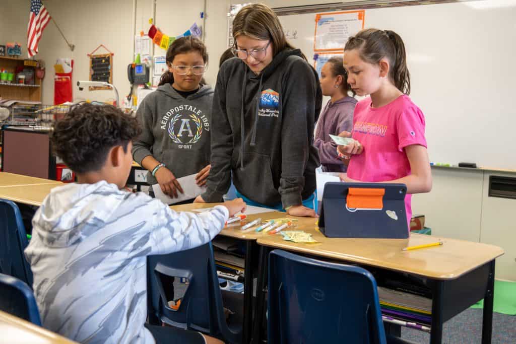 Male 5th grader sitting behind school desk with three female 5th graders in front of him looking at the markers he's selling at the class market. 