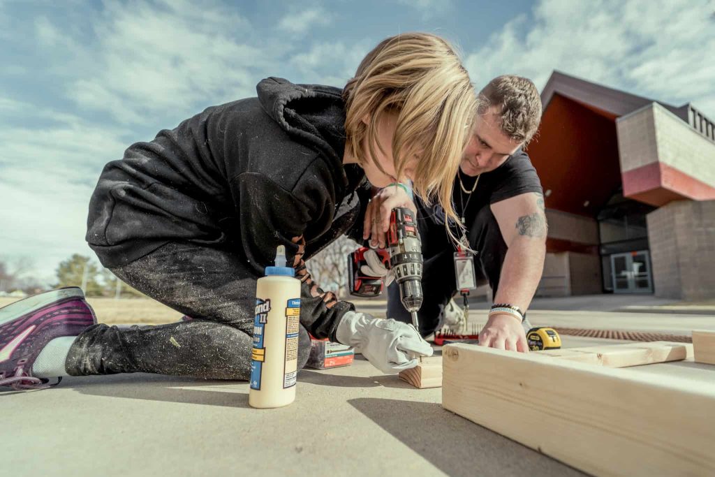 Male student and teacher on the ground with a board in front of them while they build a doghouse 