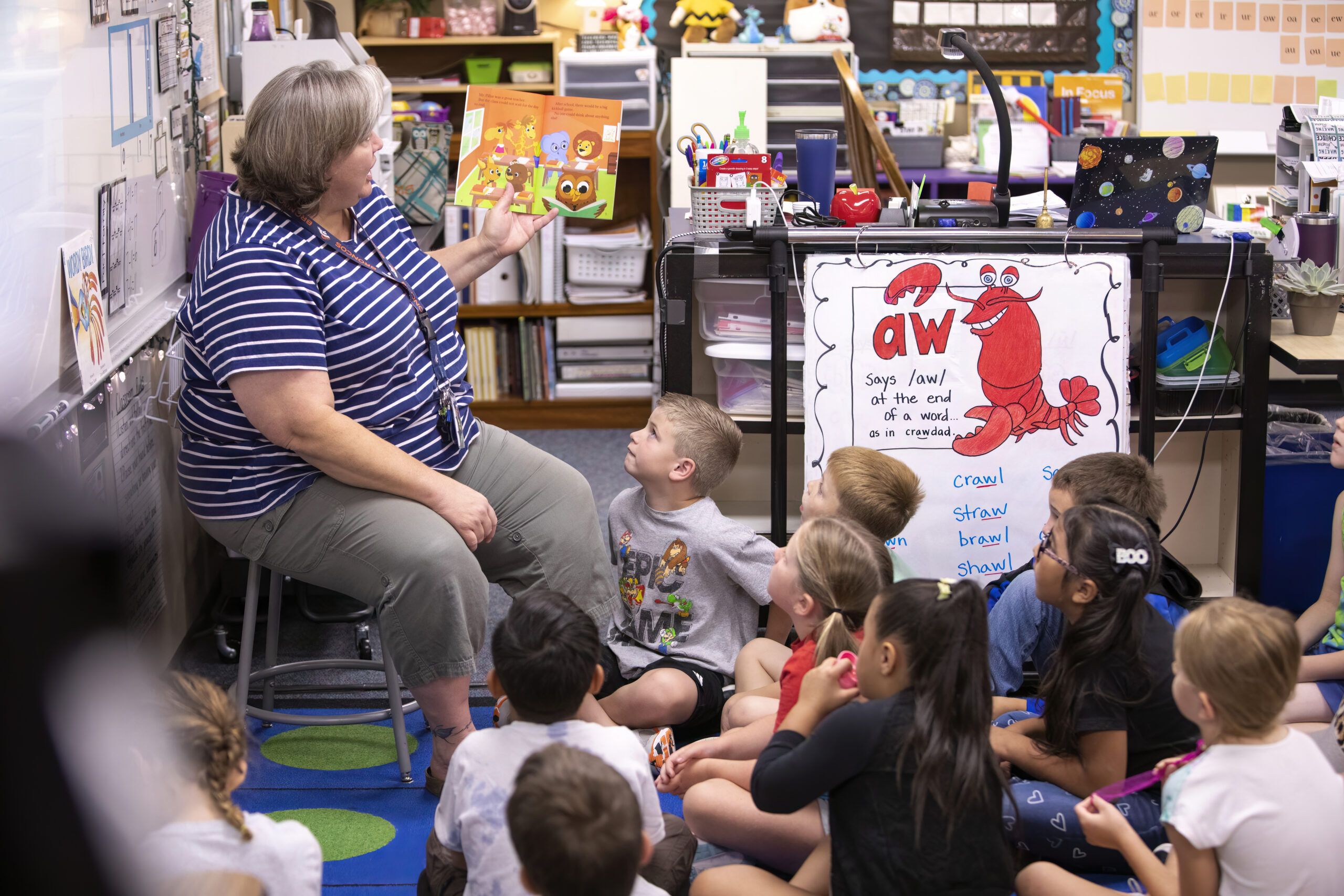 Fotografía de una profesora con camisa de rayas azules y blancas y pantalones grises, leyendo a sus alumnos.