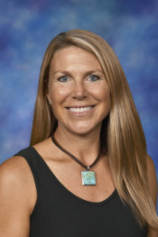 A smiling woman with long brown hair, wearing a black top and a turquoise pendant, against a blue background.