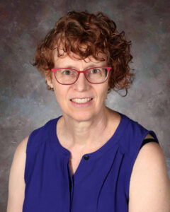 Una persona sonriente con cabello rizado, gafas y una camiseta sin mangas morada, sobre un fondo con textura.