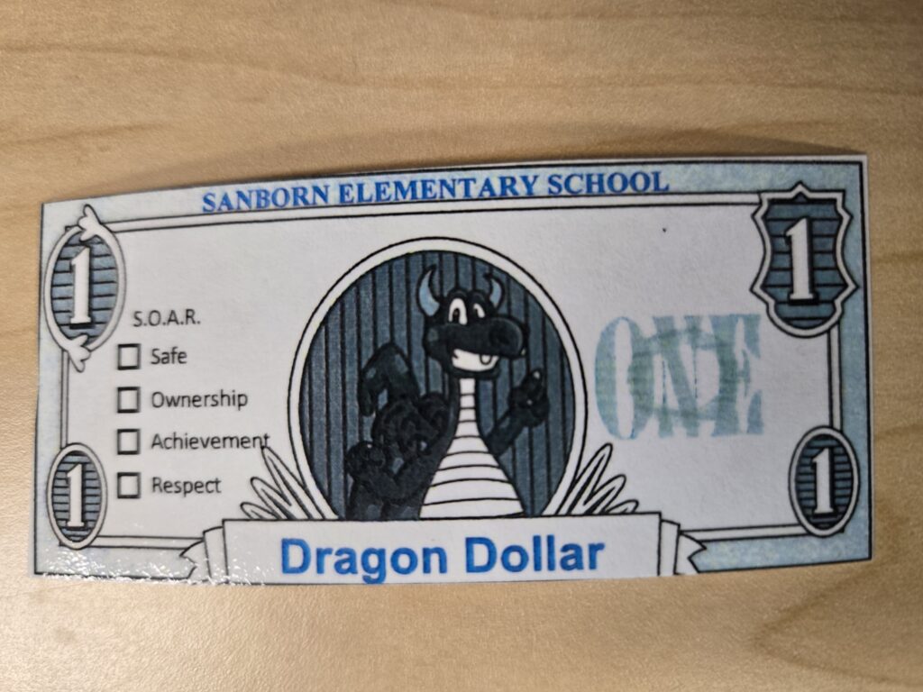 Photo of a Dragon Dollar--a fake dollar with the image of a blue dragon in the middle with the text:  SOAR for Safe, Ownership, Achievement, Respect