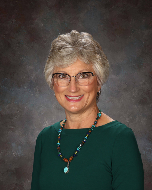 A smiling woman with short gray hair, glasses, and a colorful beaded necklace, wearing a green top against a dark background.