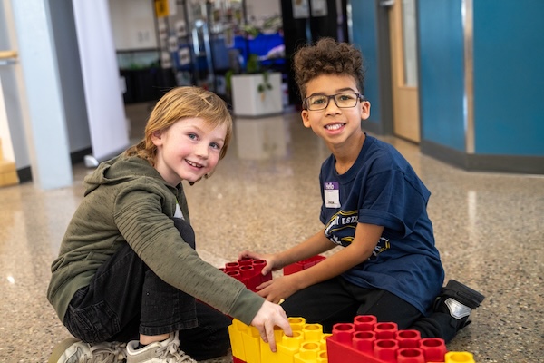 Two children are happily building with colorful blocks on a polished floor in a bright indoor space.