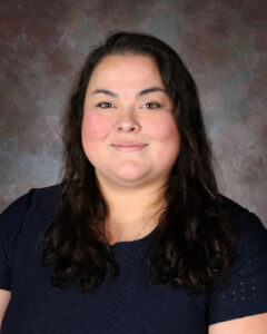 A smiling woman with long, dark, wavy hair, wearing a navy blue shirt, against a textured background.