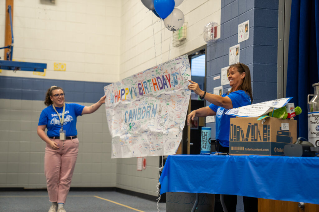 Two women celebrate a birthday in a gym, holding a colorful banner and surrounded by blue balloons and decorations.