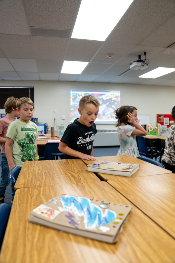 Children in a classroom interact while examining art projects on wooden tables, with a colorful projector screen in the background.
