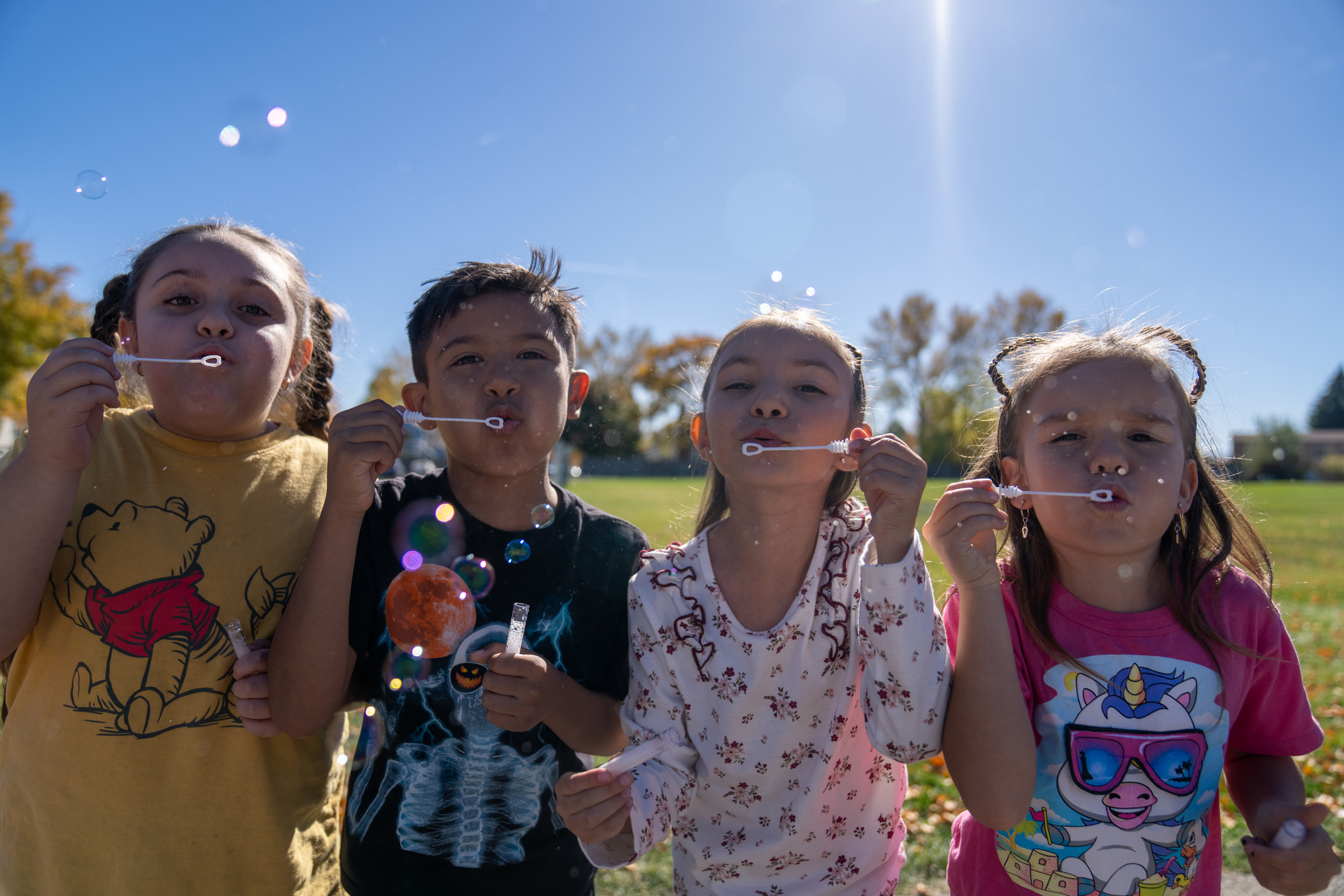 Four children blowing bubbles outdoors on a sunny day, surrounded by colorful autumn leaves and a clear blue sky.
