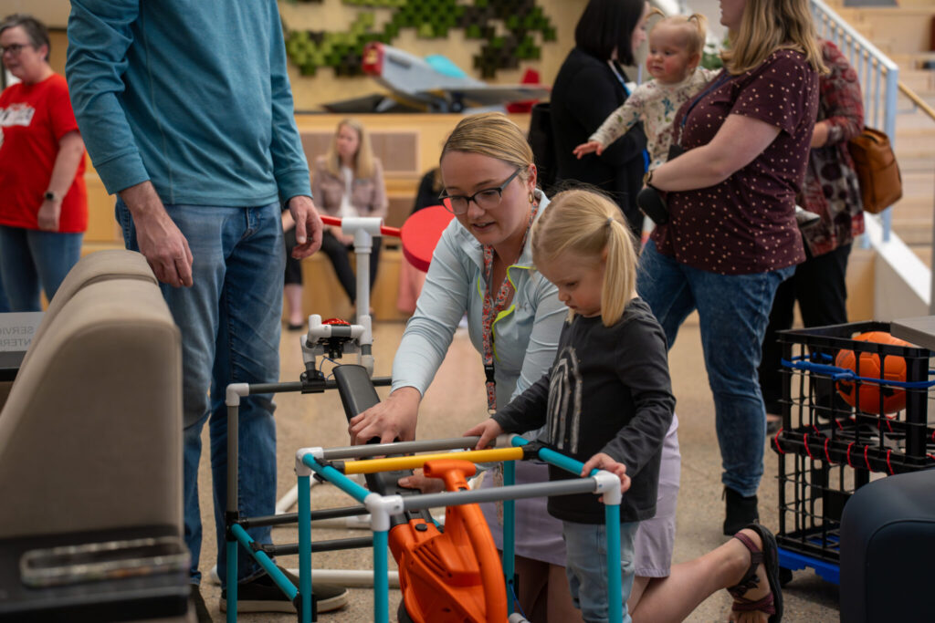 A woman assists a young girl with a colorful, adjustable tool while others observe in a busy indoor setting.