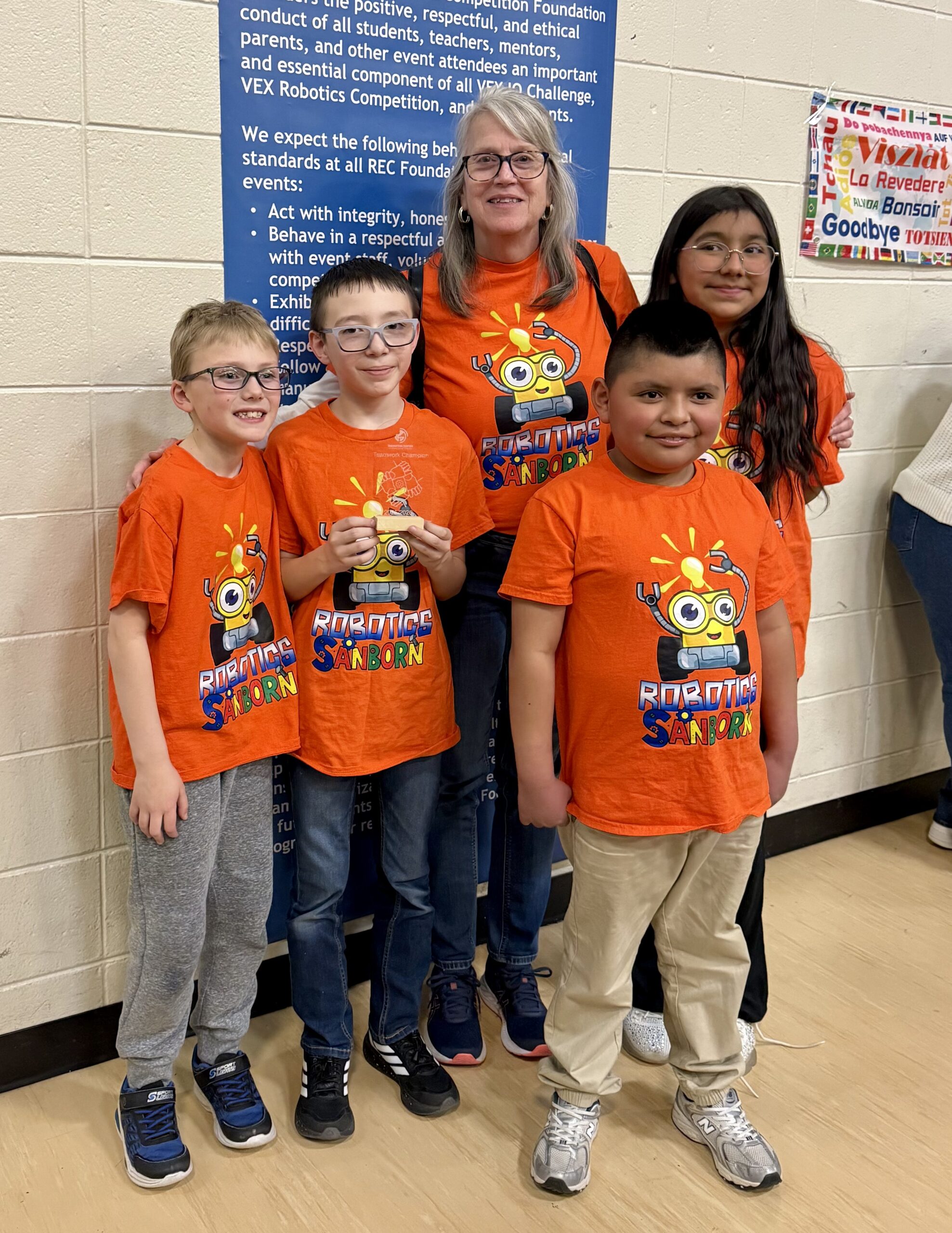 A group of five children and an adult, all wearing bright orange "Robotics Sandborn" shirts, pose together.