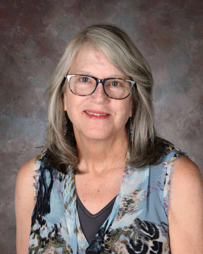 A woman with gray hair and glasses smiles, wearing a colorful sleeveless top against a neutral background.