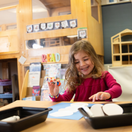 A young girl with curly hair smiles while crafting with paper and glue in a colorful classroom setting.